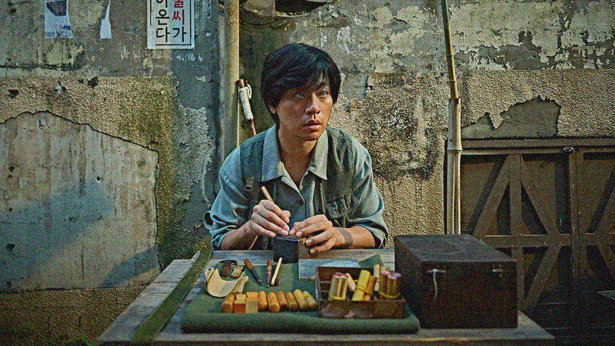 A blind man sits at a table working with tools but gazing off-screen in front of concrete walls.