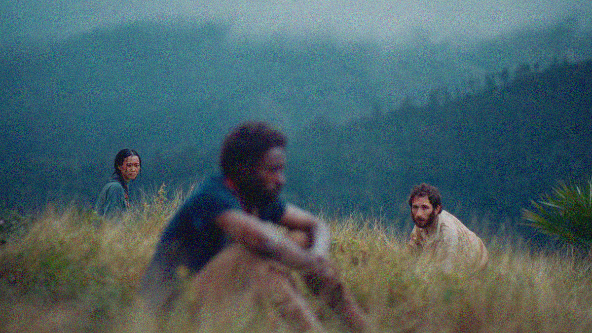 Three people sit in tall grass at the top of a hill with more in the foggy background. One man is in the middle foreground blurred so the woman at his left and man at his right are crisply focused looking at him.