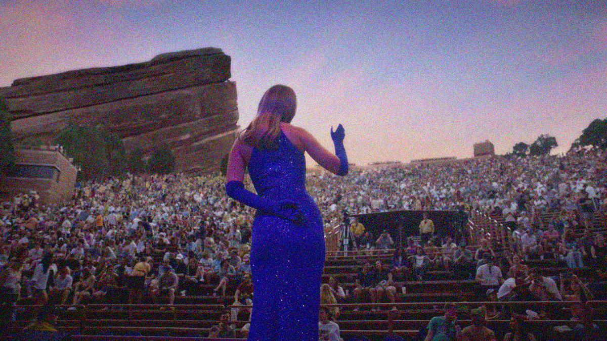 Molly Lewis in a blue sequin dress shot from behind, whistling to a large crowd on bleachers outside.