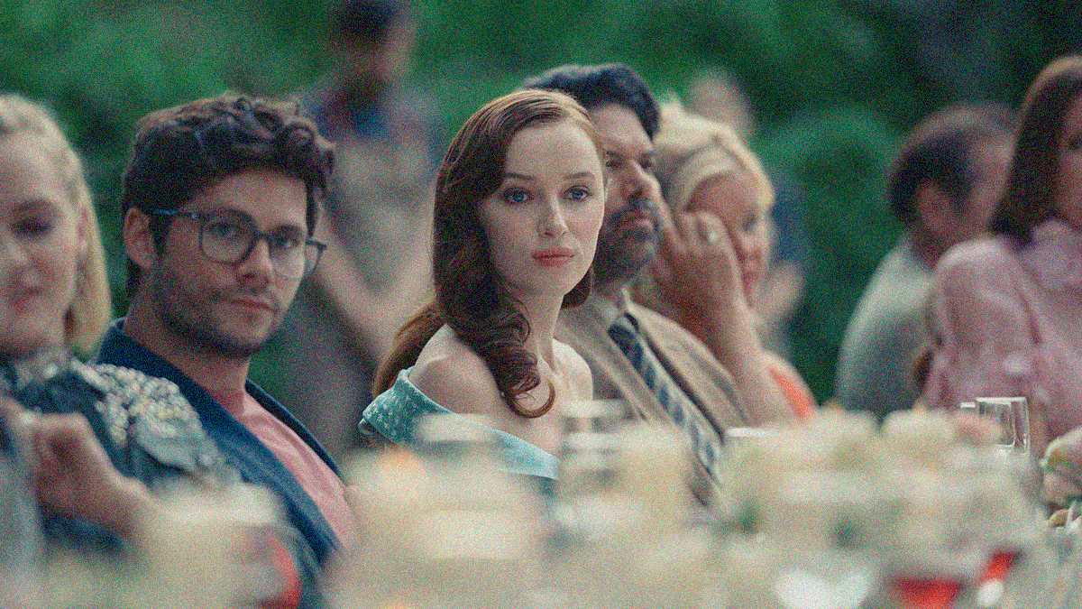A woman and man face the camera while looking just off to the right, both are seated at an outdoor dining table with many people to the right and left.