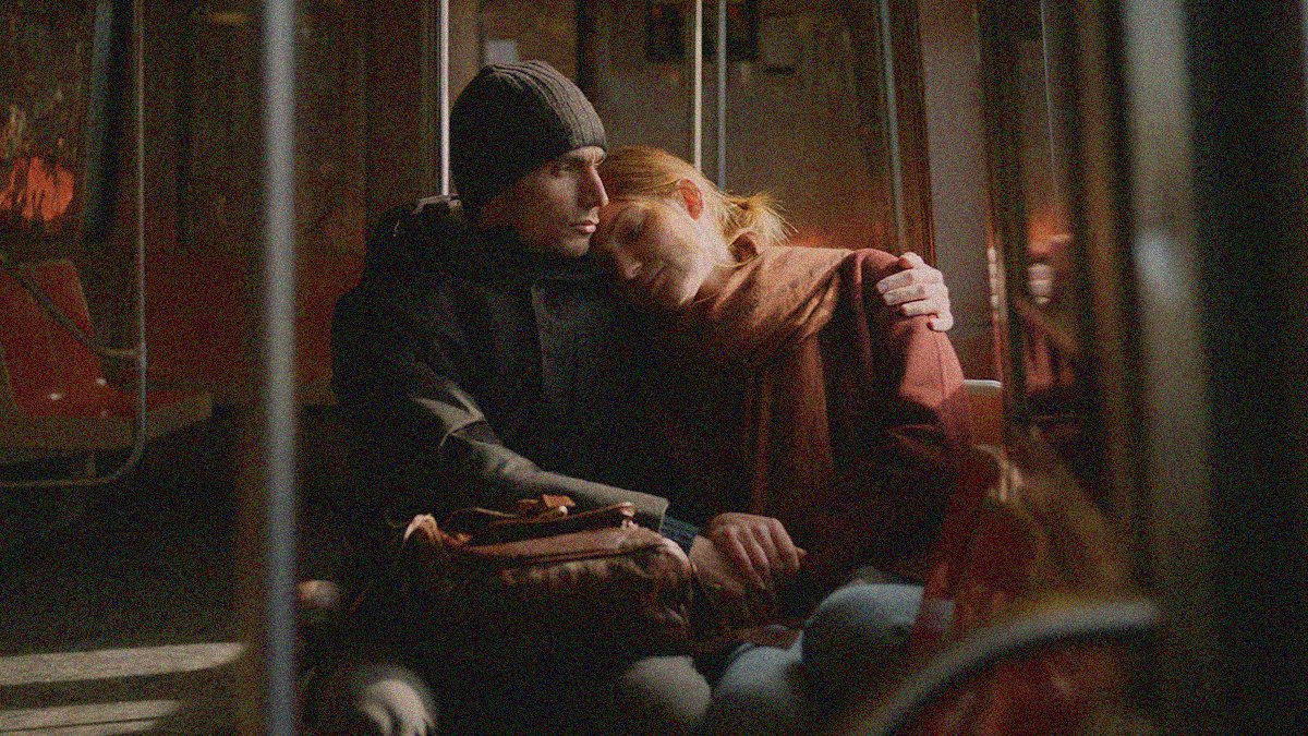 A man and a woman sitting on a subway car train bench. He looks off to the right with arm around her as her head rests on his shoulder.