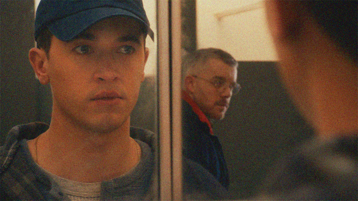 Two men are seen in reflection of a mirror. The one in the foreground is looking at the other. We stand at his back to see a bit of the real him too.