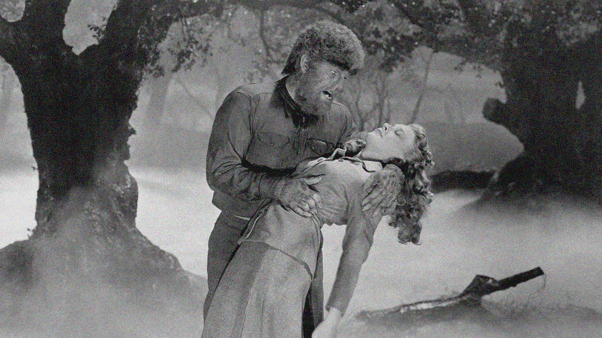 Black and white in a foggy forest, a wolf man stands with an unconscious woman bent backwards into his arms.