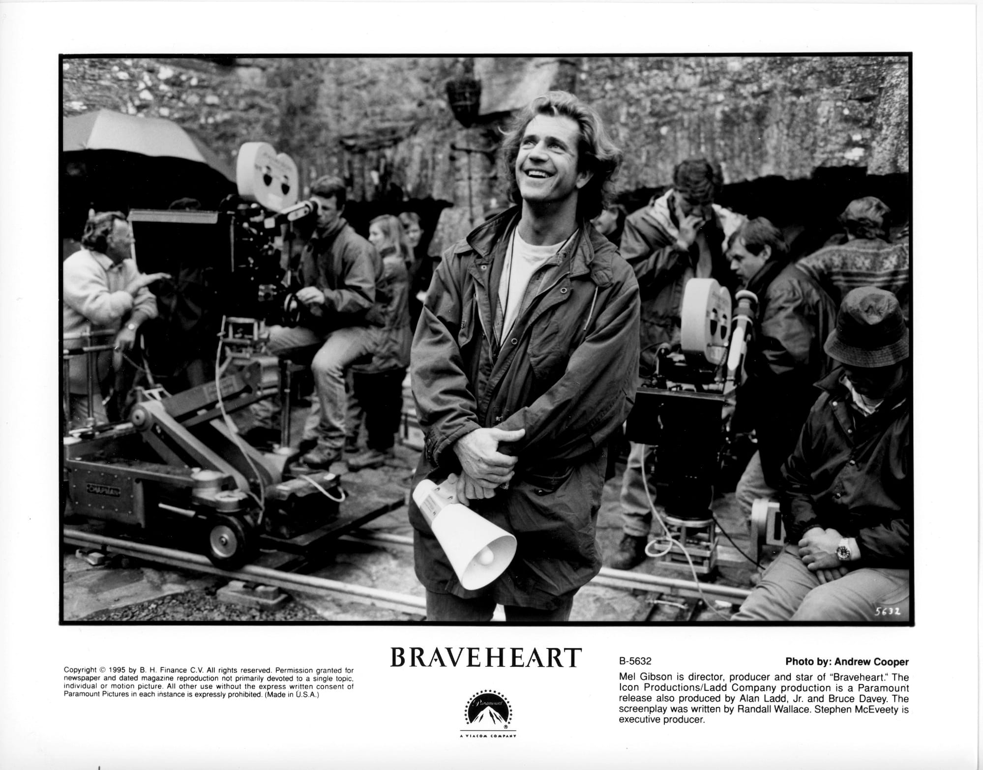 A man stands center frame smiling and holding a bullhorn while people operate two film cameras behind him.