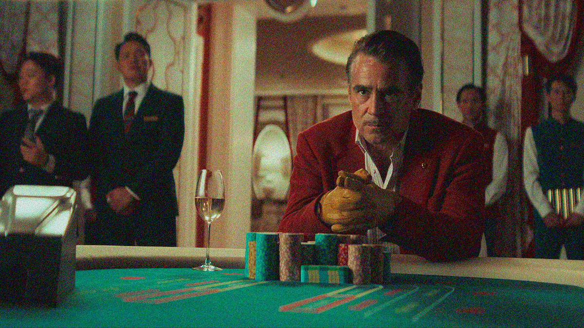 A man in a red suit coat sits at a casino baccarat table with elbows on the edge and yellow gloves clasped together.