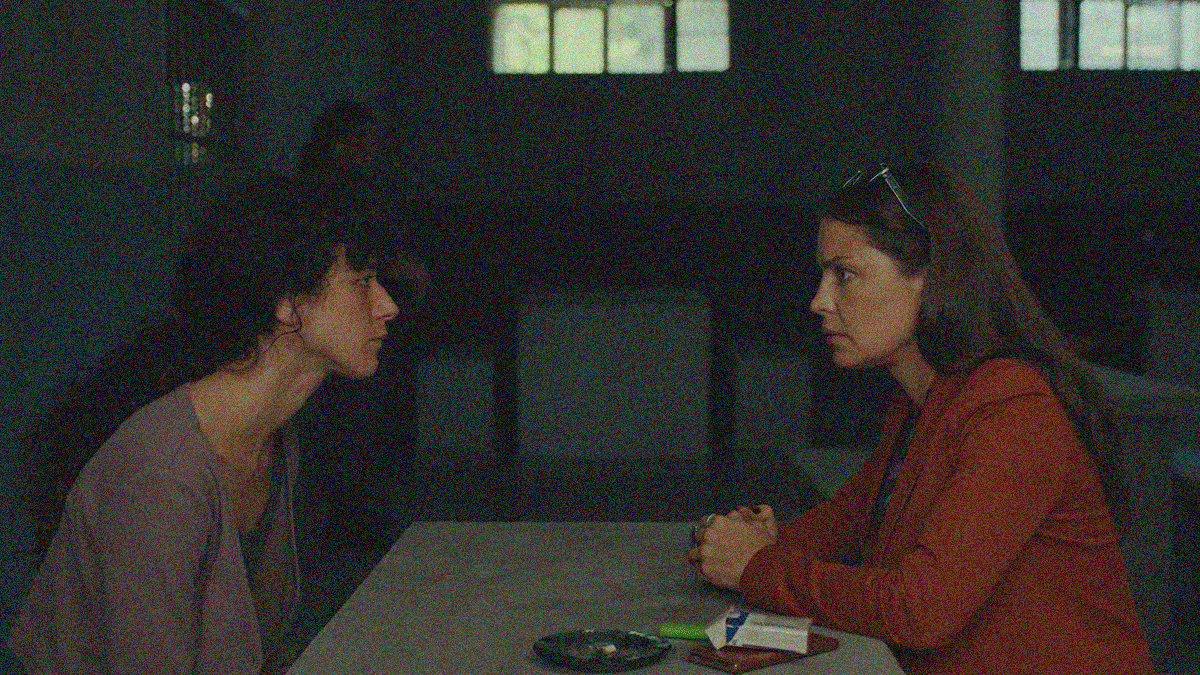 Two women sit at a prison table with an ashtray and carton of cigarettes on it. At left, a young inmate. At right, her attorney in red blazer with glasses propped on head.