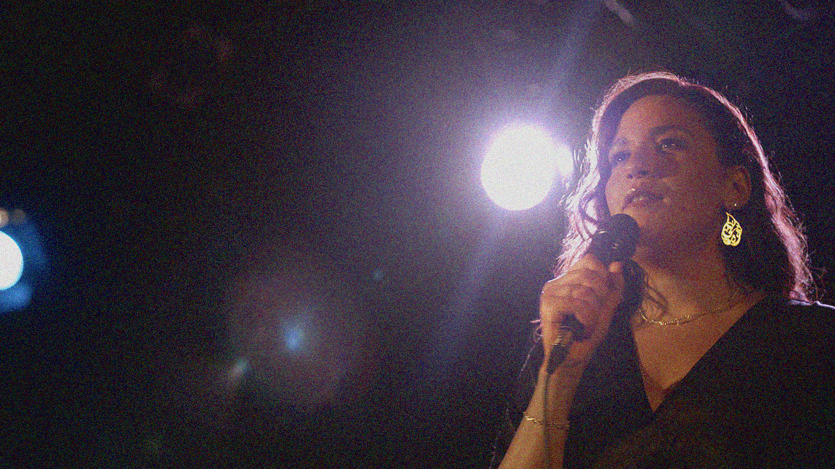 A woman is seen holding a microphone from chest up in the right side corner. The dark room highlighted by two bright stage lights fills the rest of the frame.