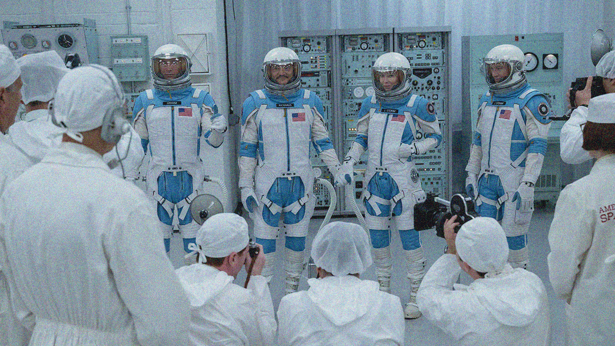 Four astronauts are side by side in white suits and helmets with baby blue highlights. Vintage computer servers are seen behind them. Photographers in white film them.