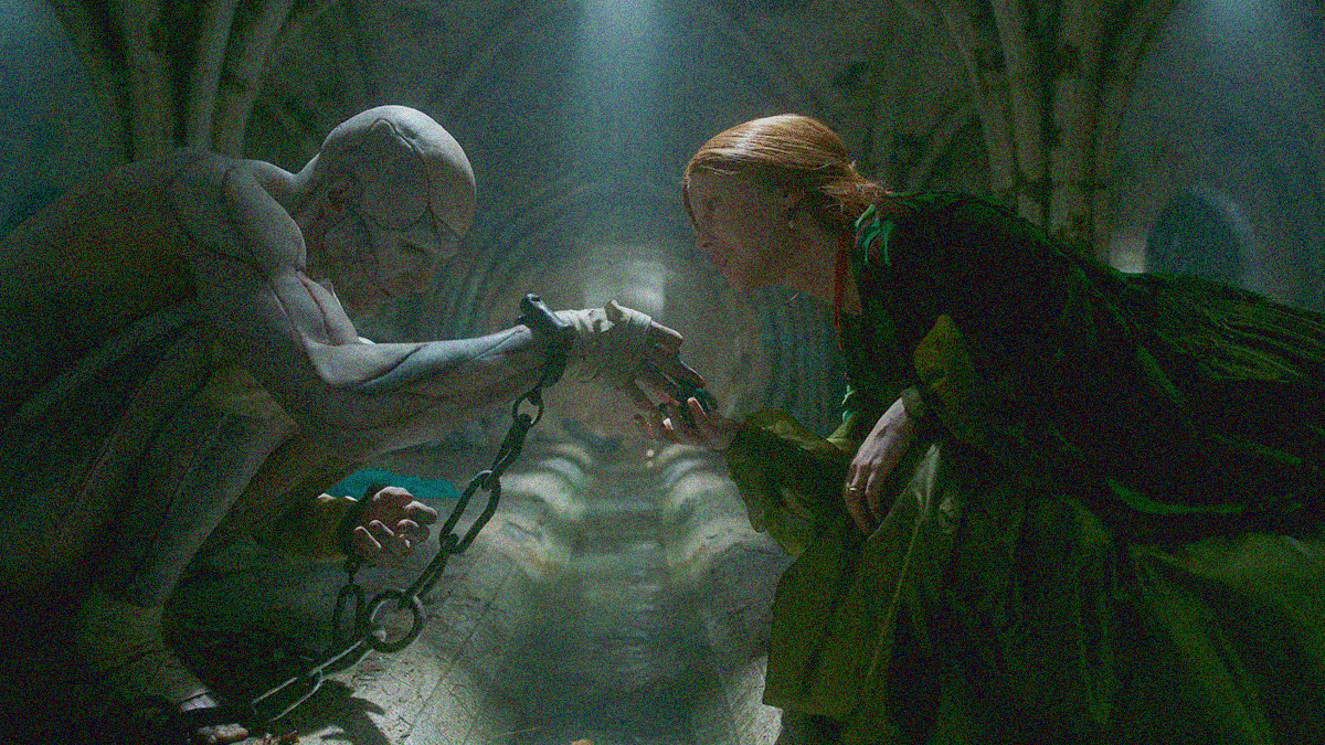 Two figures crouch down in front of a tunnel with a small stream of water between them. The man on the left is naked, covered in scars, and chained. The woman on the right is in a flowing dress, her hand reaching for his.