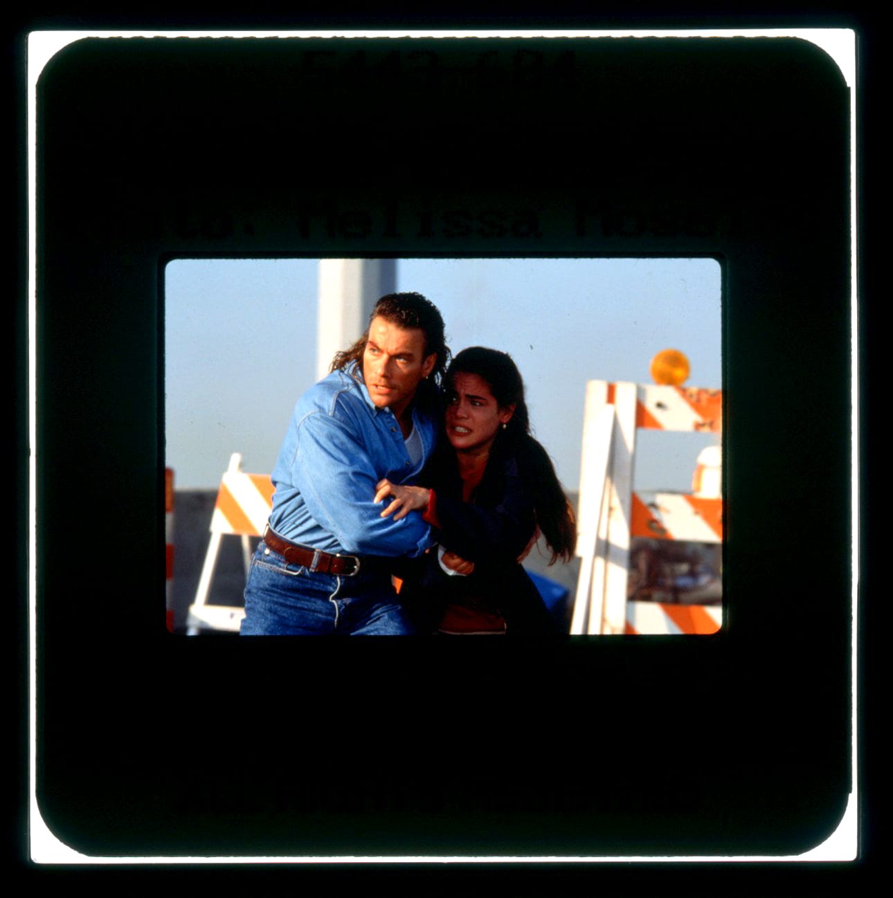 A man and woman embrace in a protective stance with him lean over her as both look off-screen left.