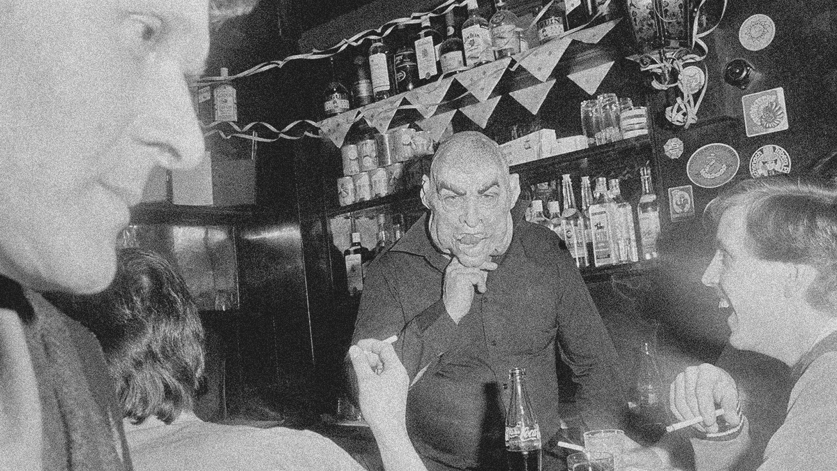 Interior of a bar in black an white. There's the large face of a man on left side coming into frame and a smaller face of a man laughing with cigarette in hand at bottom right. At center is a third man wearing a rubber Halloween mask.