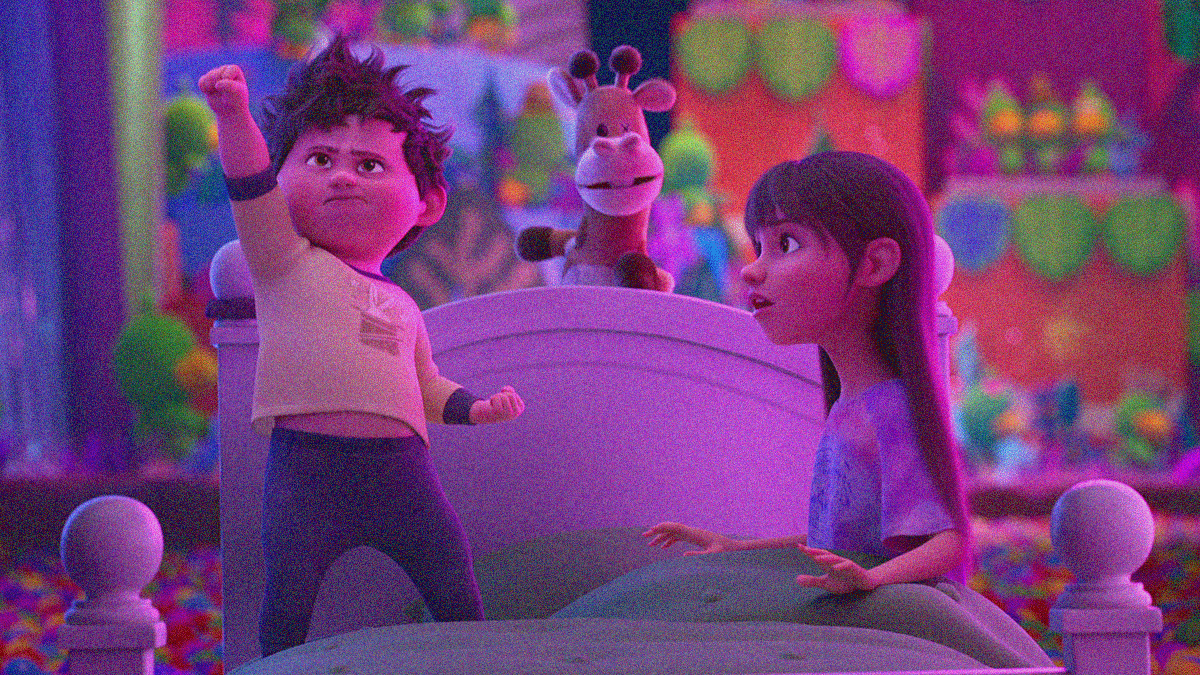 A boy stands on a bed with one fist in the air while his slightly older sister sits and looks at him. A stuffed giraffe with one eye balances on the headboard between them.