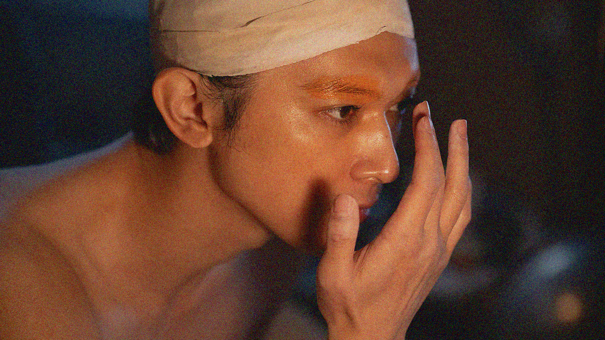 Close-up of a shirtless man in a head wrap moving his finger to apply something to his eyebrow.