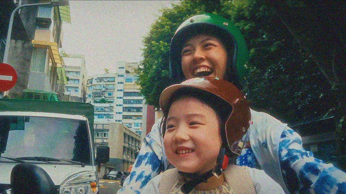 Close-up of two people (a girl sitting front of a woman) wearing helmets on a motorcycle, smiling and laughing. 