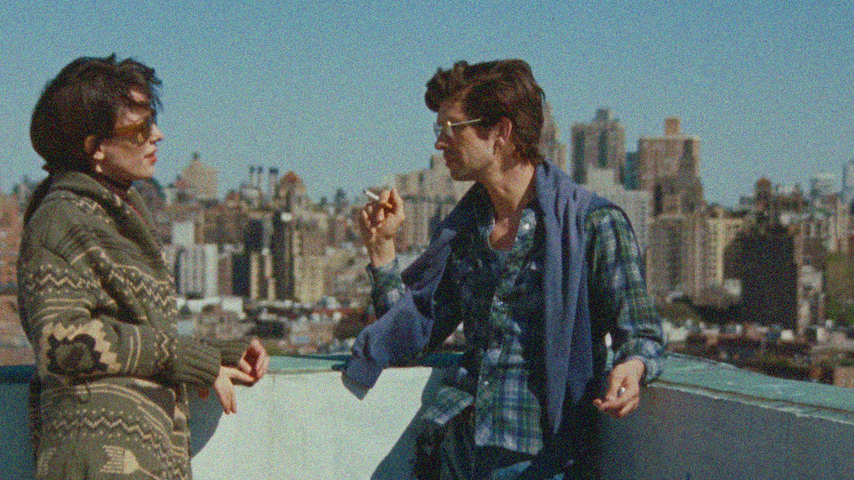 A woman in a sweater looks at a man smoking in button-down and scarf. Both lean against the edge of a balcony, New York City skyscrapers fill the space behind them.