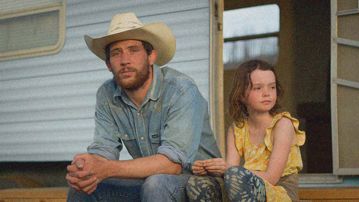 A man in a cowboy hat seats beside a young girl in yellow outside the open door to a trailer.