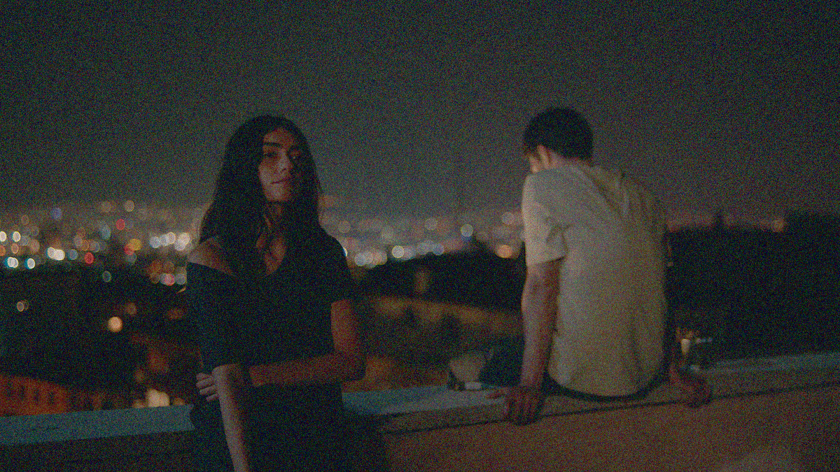A woman leans against the parapet of a roof facing the camera and a man sits atop it facing the cityscape and night sky.