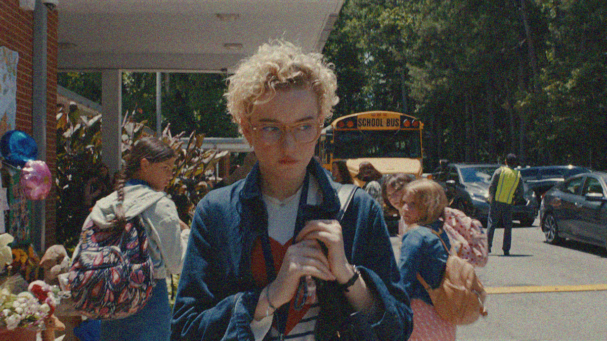 A blonde woman with glasses walks towards the camera, away from children staring at her from outside a school.