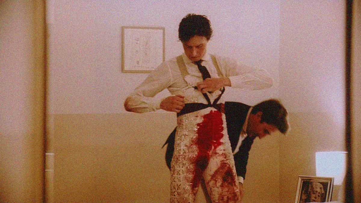 A matador in bloodied costume unties black ribbon around his waste as another man undoes his pant leg.