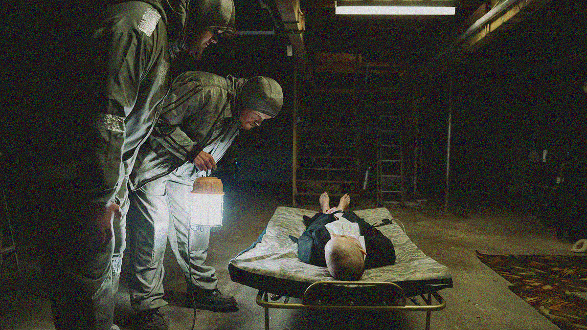 Two men in duct taped faux hazmat suits bend down to peer at a bald, unconscious woman lying on a futon mattress in a dark basement..