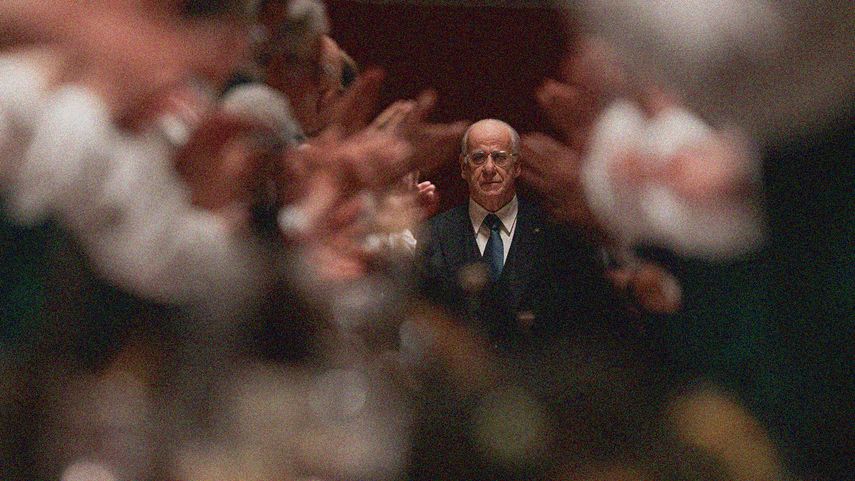 A balding older man in a suit with glasses can be seen between the blurred visages of people standing and clapping on the left and right of him.