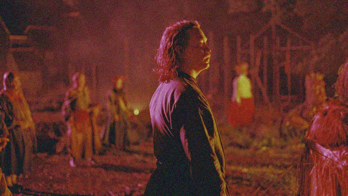 A man with long hair stands center frame looking right as the people, fields, and property behind him are awash in the reflected red of flames.