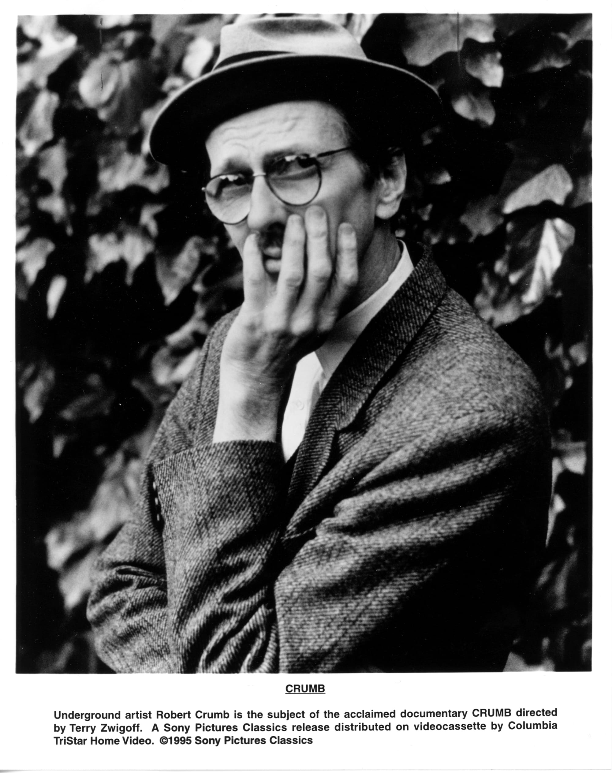B&W publicity photo: Crumb shown from waste up in glasses and hat, right arm folded beneath left elbow and left hand supporting his head and covering his face so a part of his mouth and mustache is seen between his pinky and ring fingers.