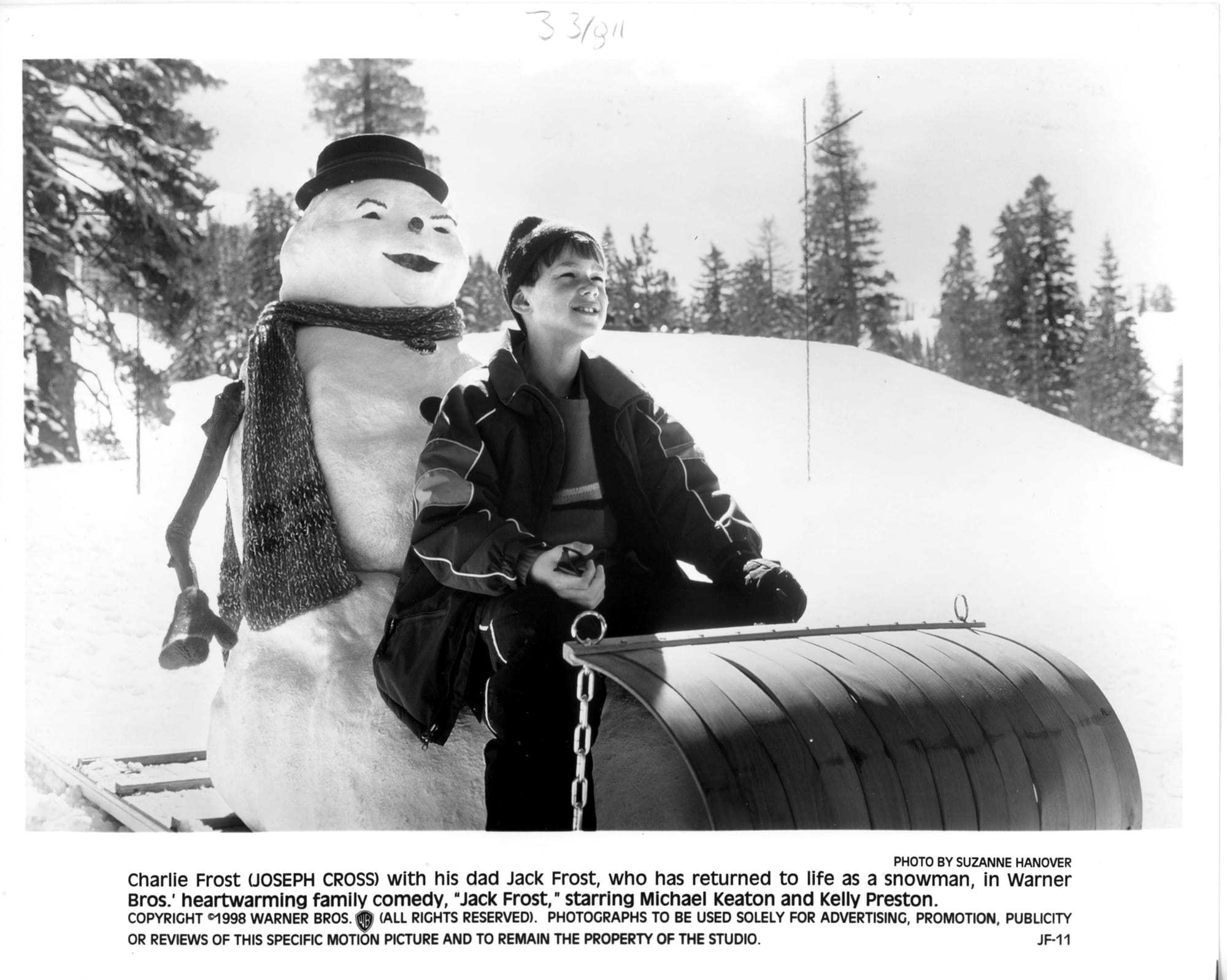 B&W Publicity photo: A snowman stands behind a boy on a giant sled as they go down a snowy hill smiling.