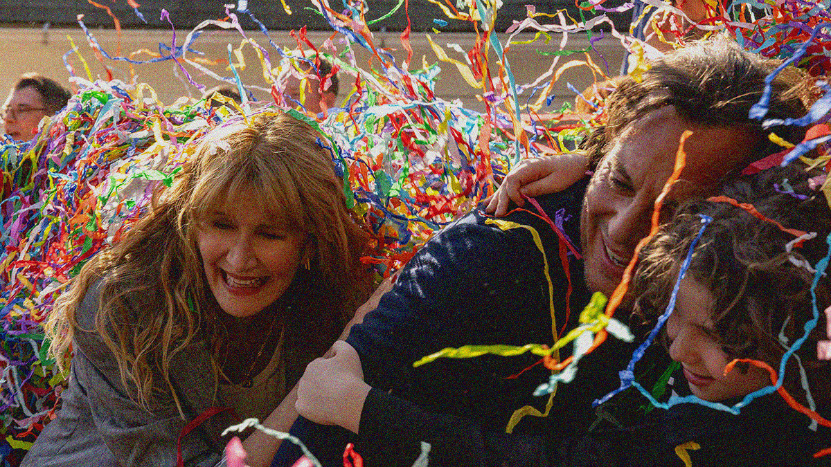 A woman leans over while laughing onto a man's back as he laughs and falls onto their son in an embrace while colorful confetti streamers fill the screen.