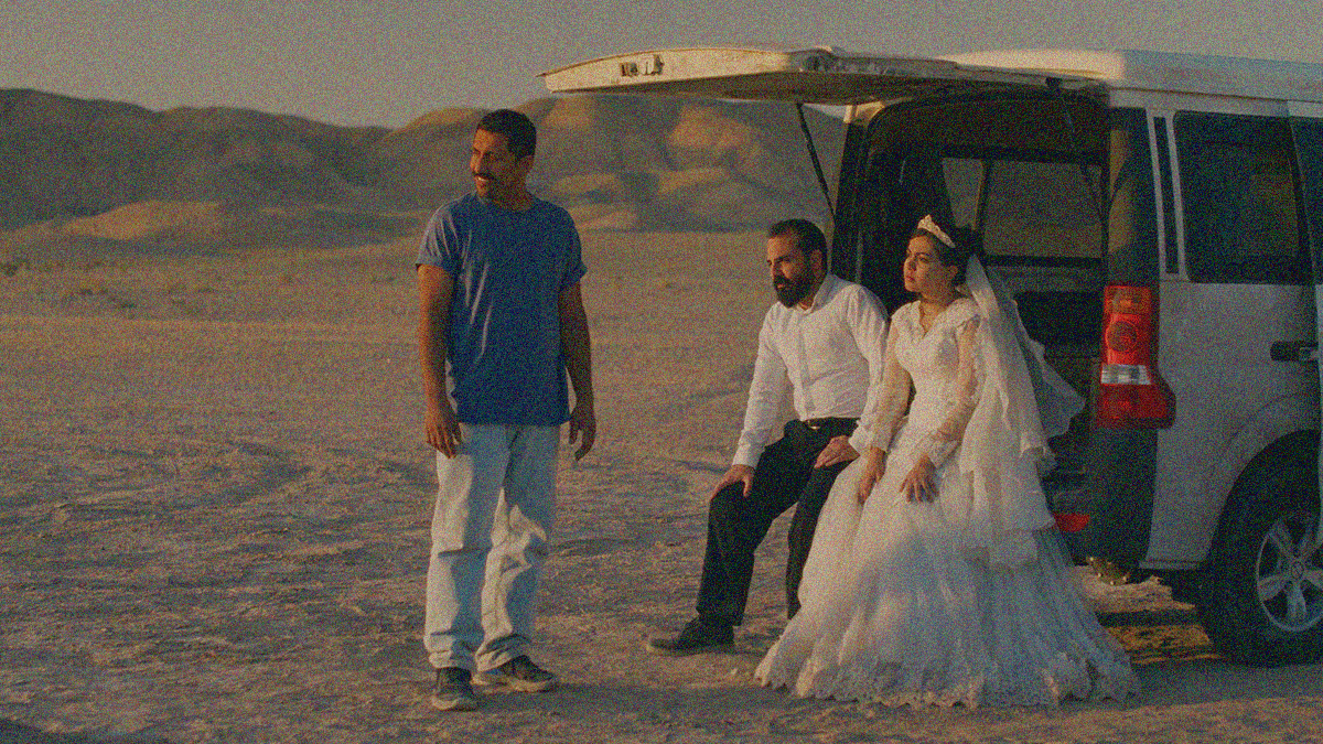 A man in blue t-shirt stands and smiles off-screen to the left while a bride and groom sit on the bumper of a van with the trunk door open.