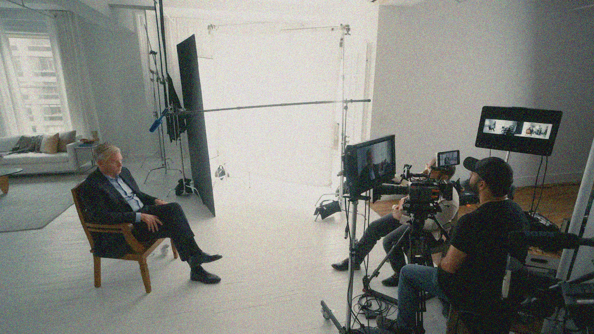 A man in a suit coat sits in a chair at left while two others sit in front of and behind a camera set-up to film and interview him at right.