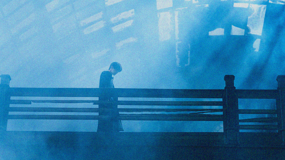 A man is seen walking left to right on what looks to be a bridge. The ceiling above is full of broken windows allowing the blue-tinted light to diffuse through.