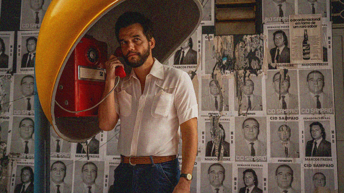 A bearded man stands with a red pay phone receiver against his ear beneath a yellow shell enclosure and in front of a wall plastered with B&W photos of men.