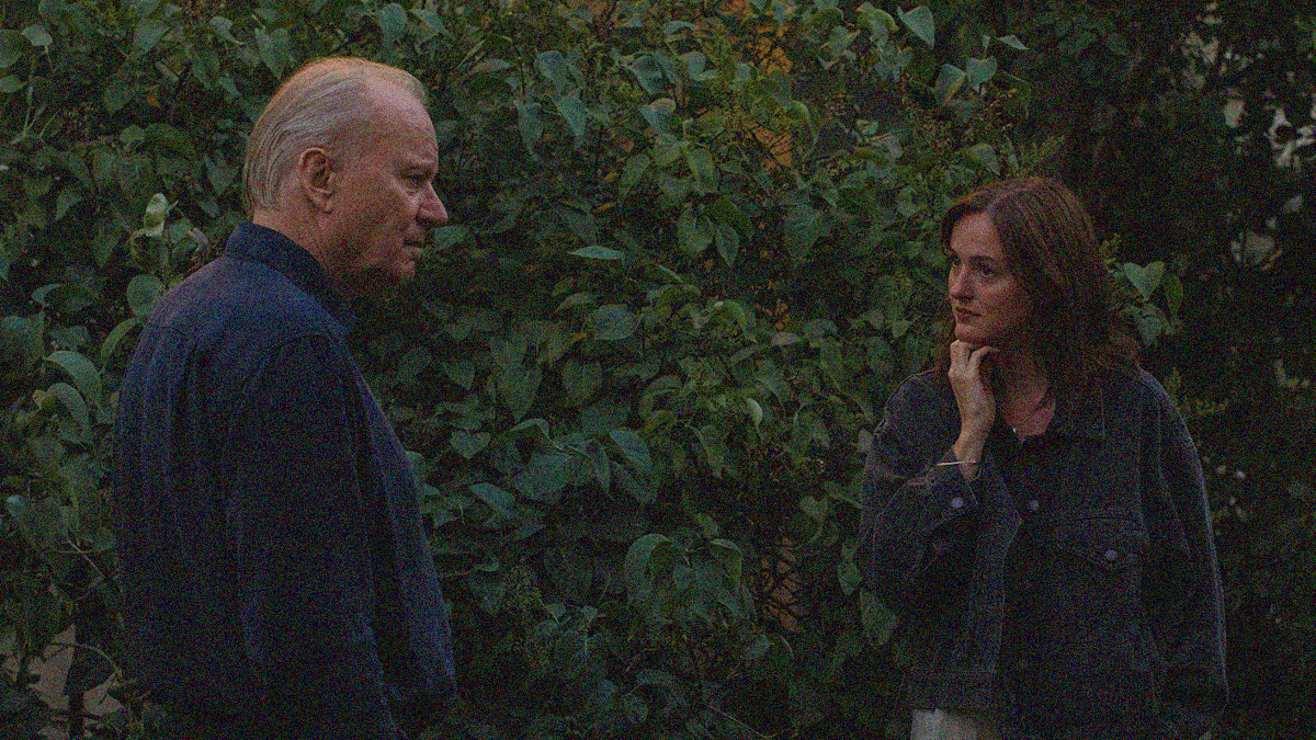 An older man stands at left looking into the distance while a young woman stands at right looking at him. They stand against a wall of leaves.