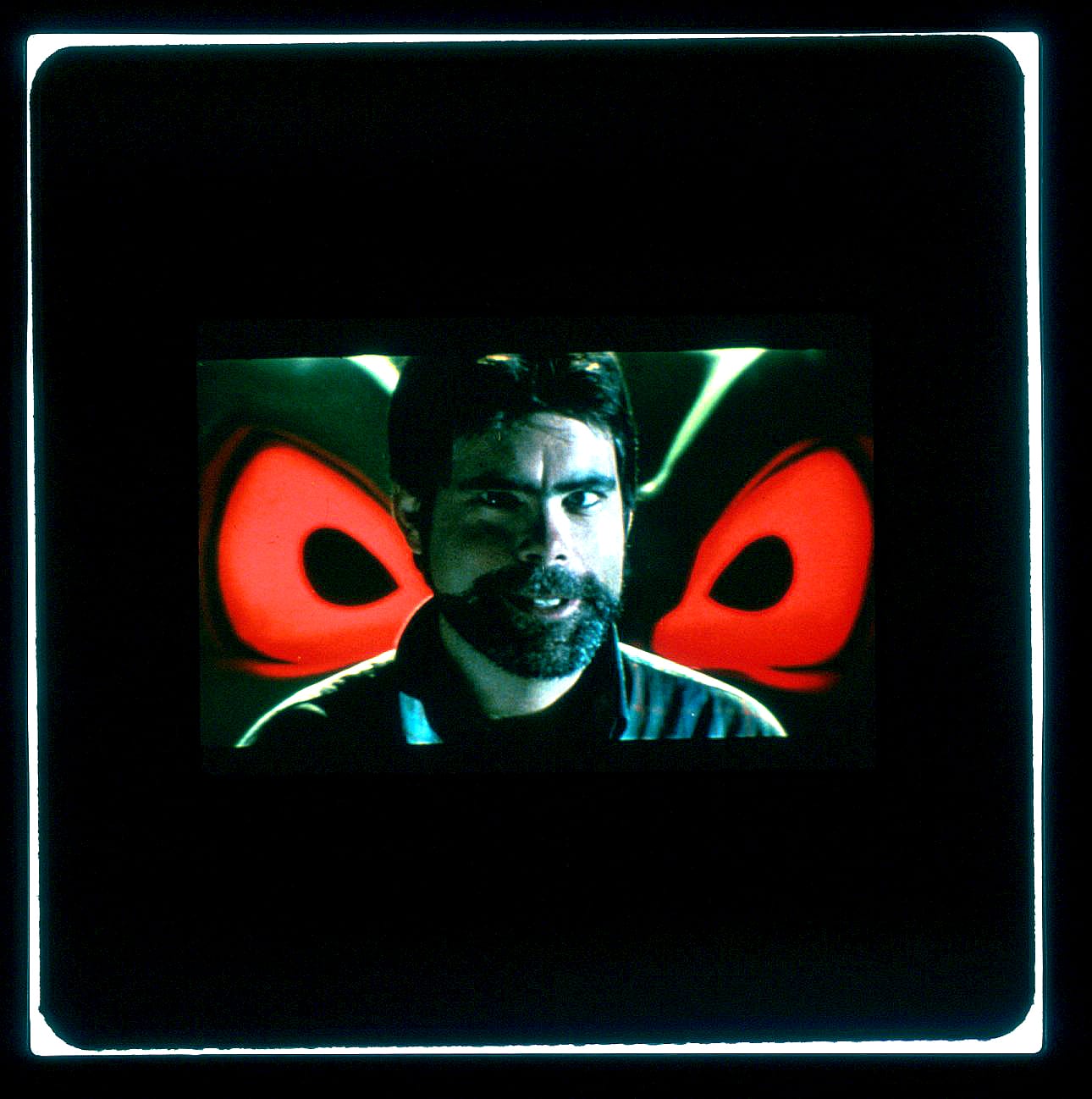 Color Publicity Slide: A bearded King from shoulders up smiles into camera between the glowing red eyes of the demon truck behind him.