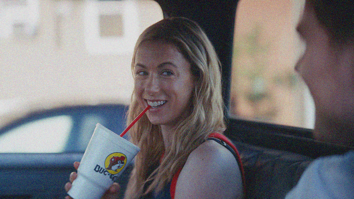 A blonde woman in center frame, smiling with a red straw in her mouth (from a large white plastic cup), looks right at a blurred man's face in foreground.