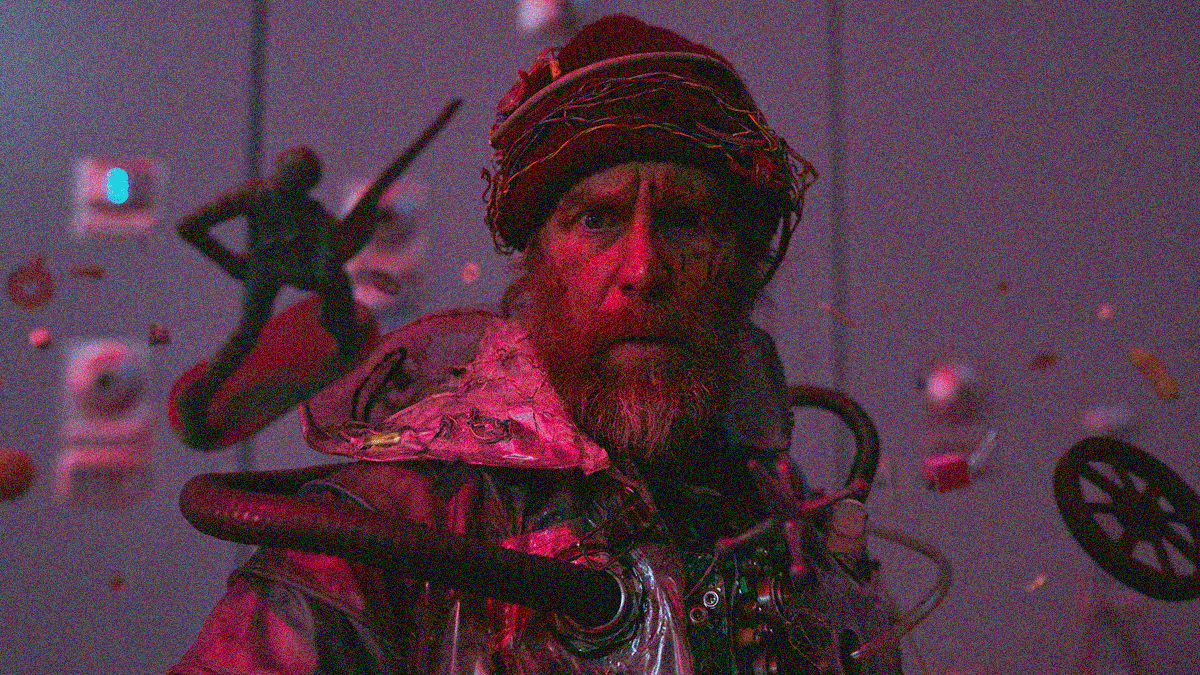 A bearded and disheveled man in a plastic hooded coat and wire-wound winter hat stares in the camera as objects (an army man and wheel) float around him.