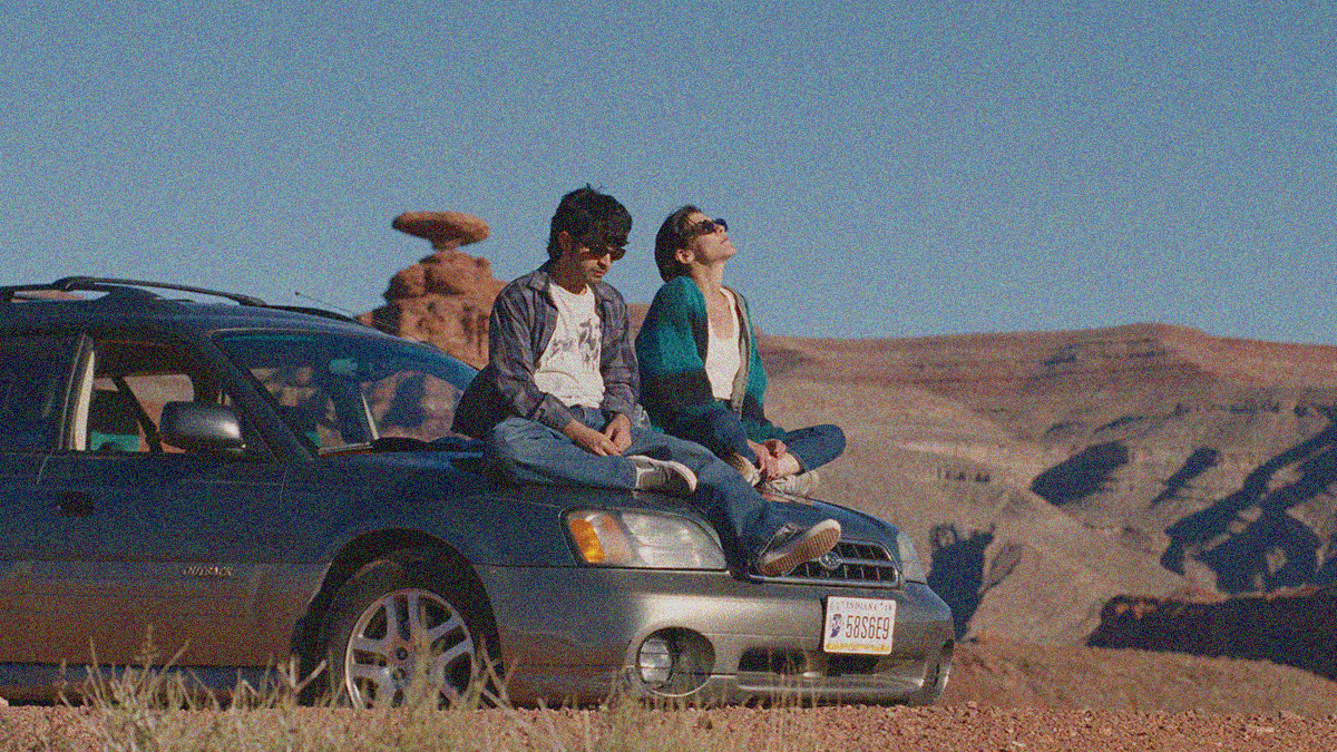 A man and a woman sit on the hood of a Subaru Outback in the desert. Both wear sunglasses. He looks down. She looks up.