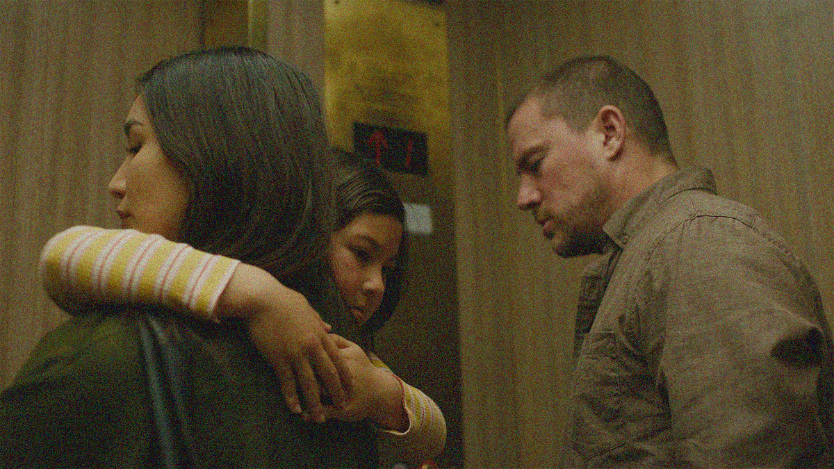 A woman at left has a young girl wrapped around her neck. The former looks left, the latter looks right over her shoulder. A man stands next to them at right. All three are in front of an elevator, waiting.