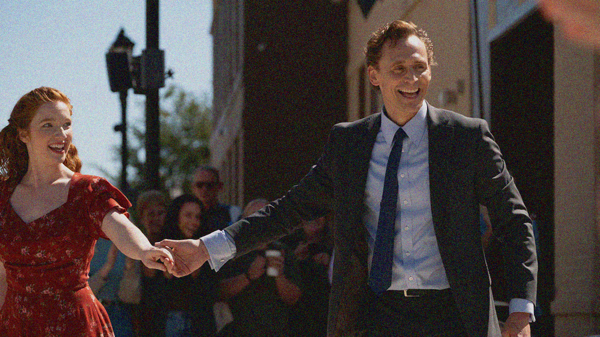 A man in a suit and tie smiles at the right of the frame, his hand pulling a woman in a red dress into frame from the left.