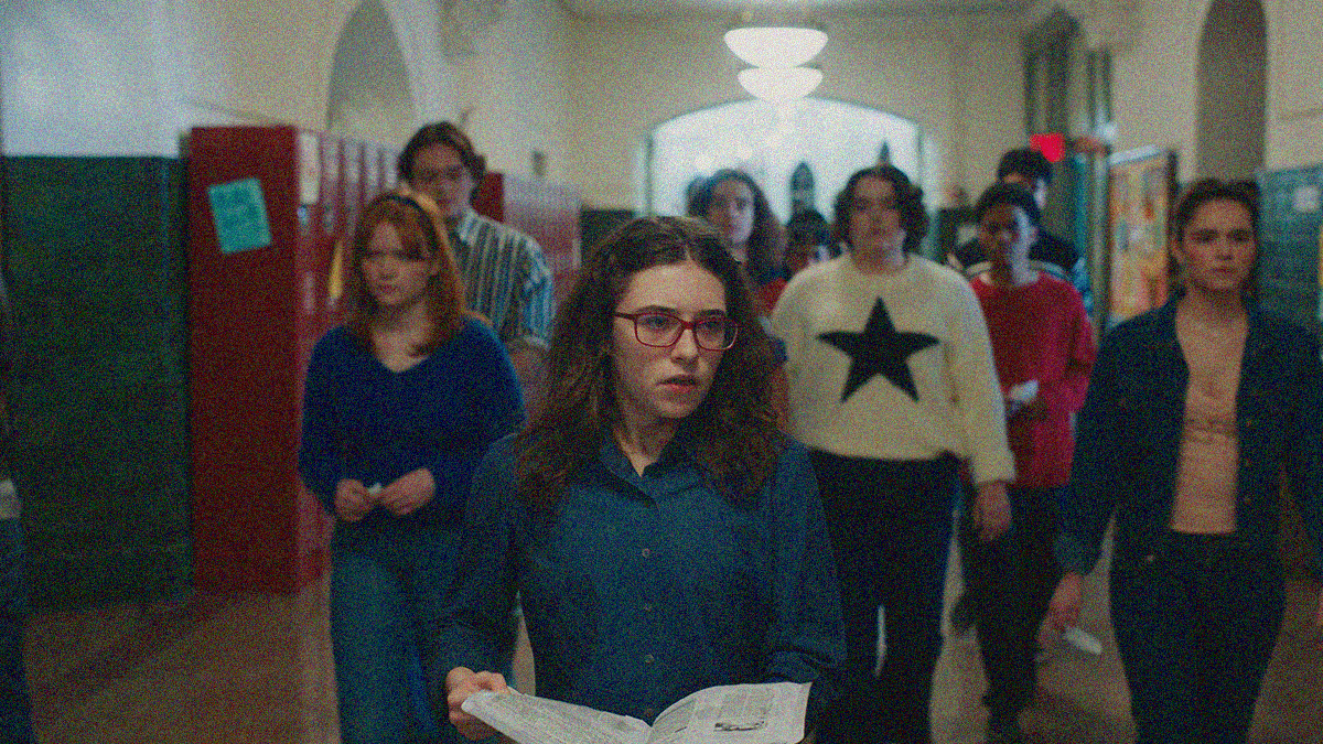 A teen girl stands front and center with glasses, an opened book in-hand. A school hallway with red lockers and a group of other teens is seen behind her.