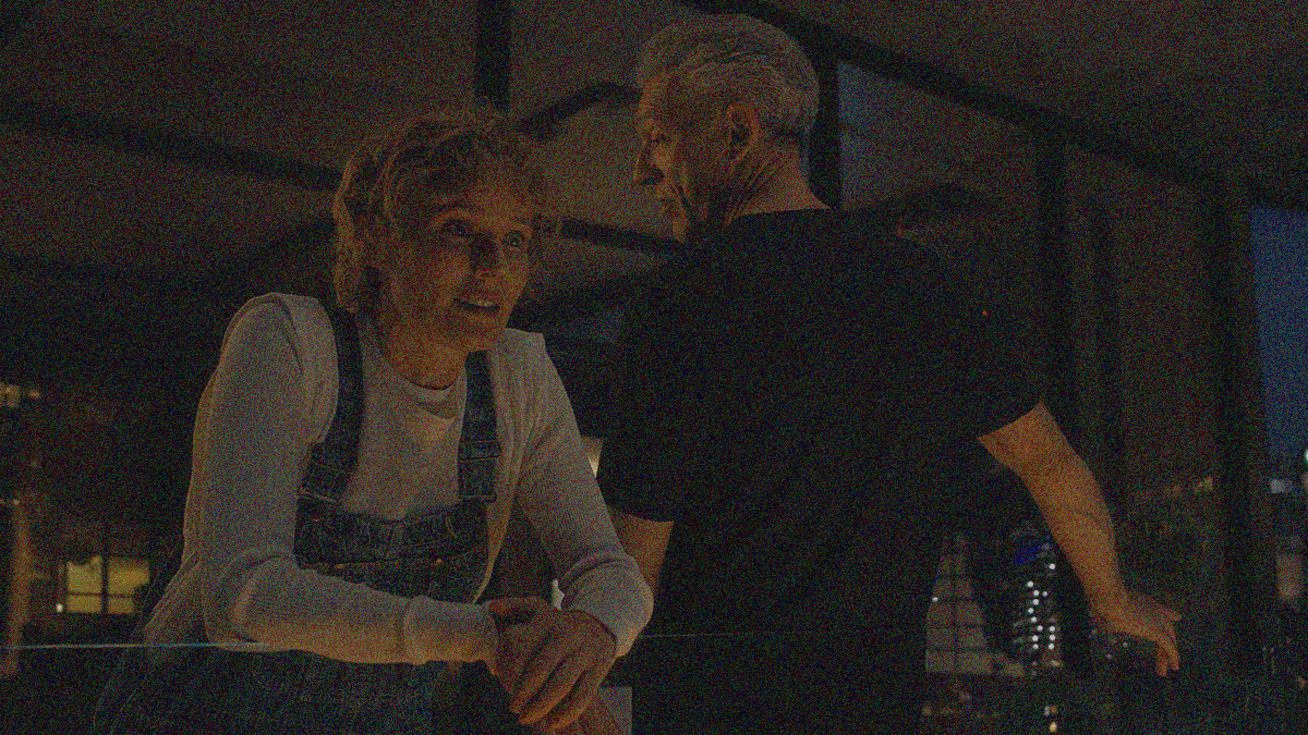 A woman and a man lean against the glass half-wall acting as a balcony railing. Her elbows rest on it as she looks out towards us. His lower back and right hand touch it as he faces his apartment, head slightly turned to look at her.