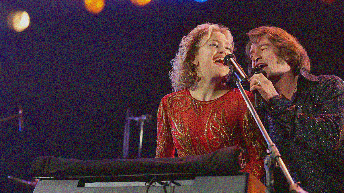 A woman in red sequins and man in black sequins are seen in the bottom right corner singing above a keyboard on a darkened stage.