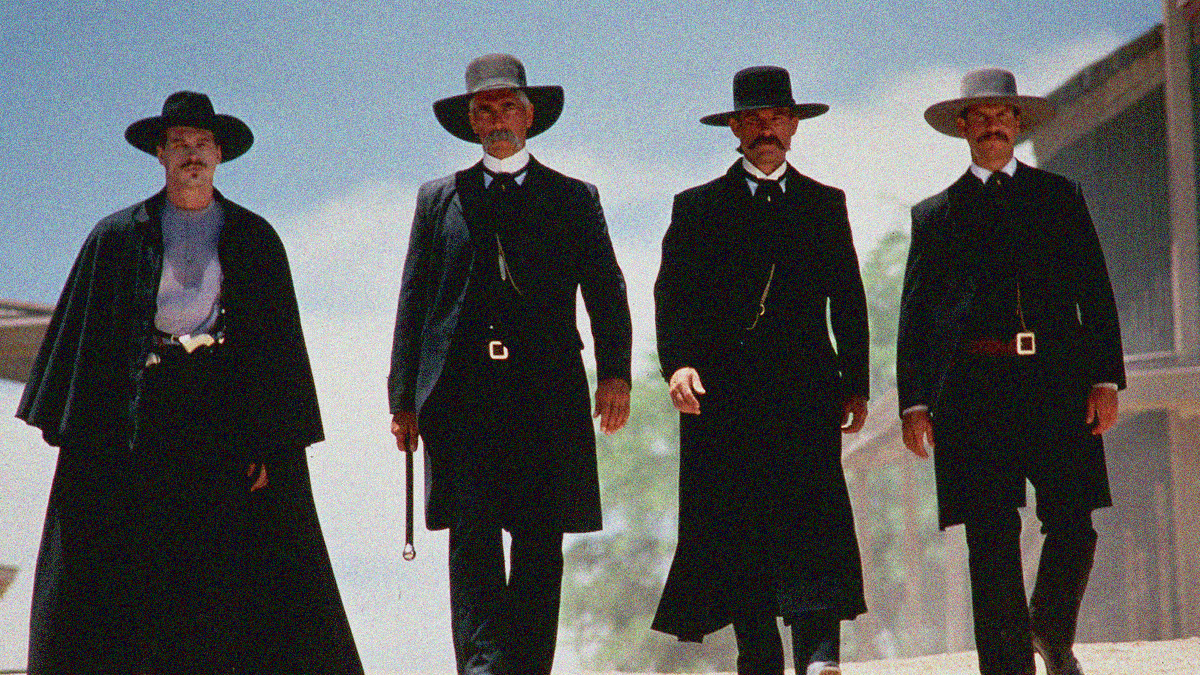 Four mustached men in all black suits and cowboy hats walk confidently towards the camera.