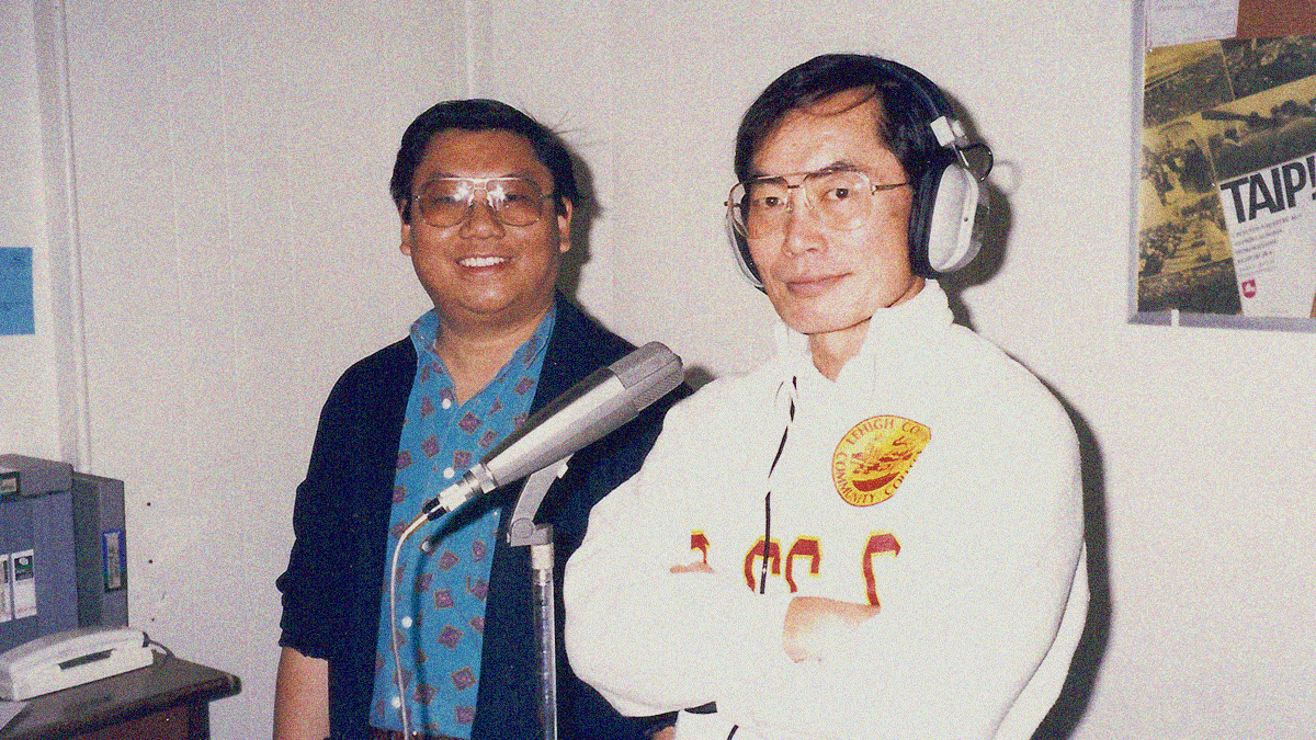 Two men stand behind a microphone. One to the side, another with headphones and arms crossed. Both smile and look into the camera.