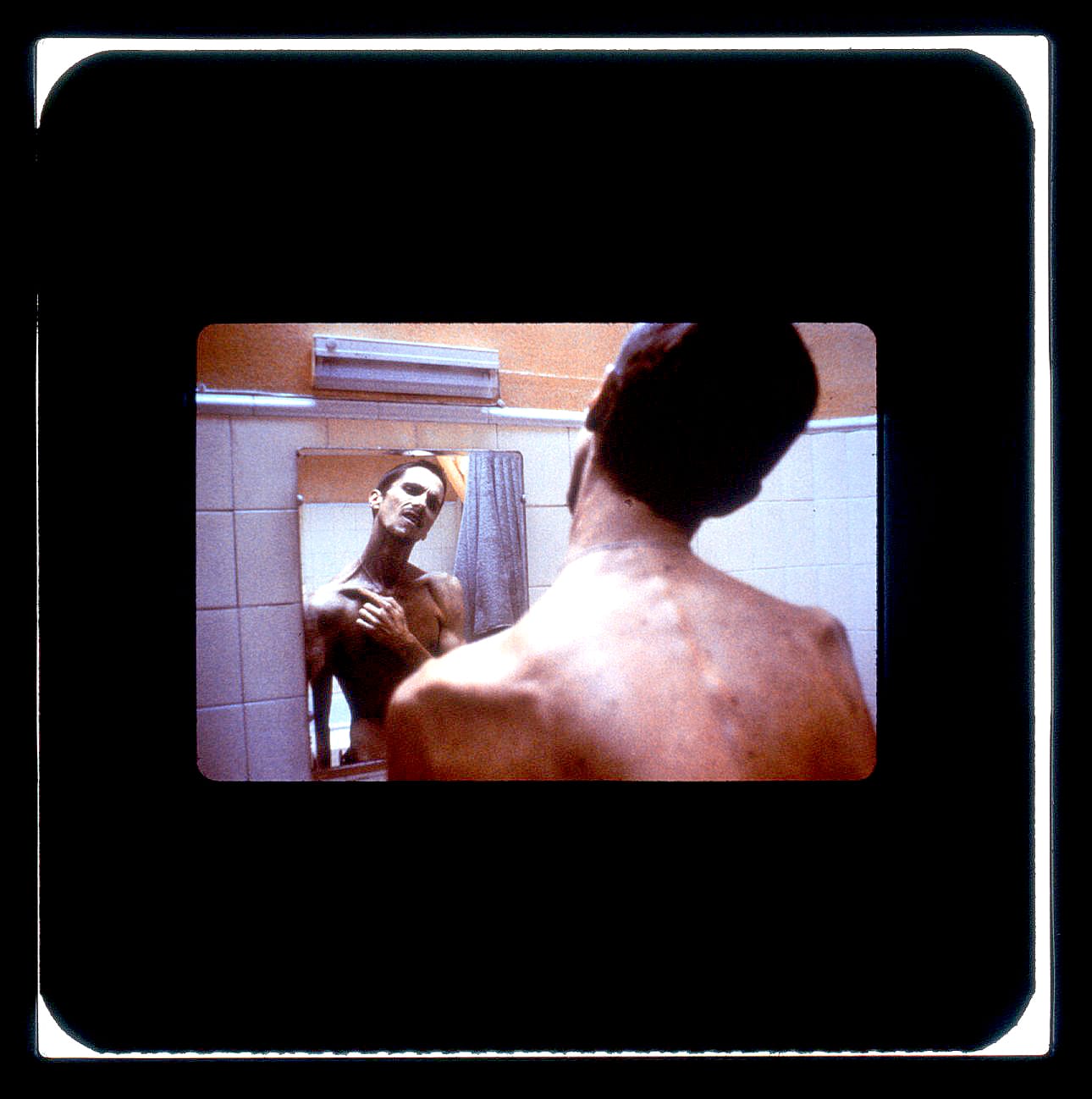 An emaciated man is seen from behind looking into a bathroom mirror. We see him touching his clavicle in the reflection.