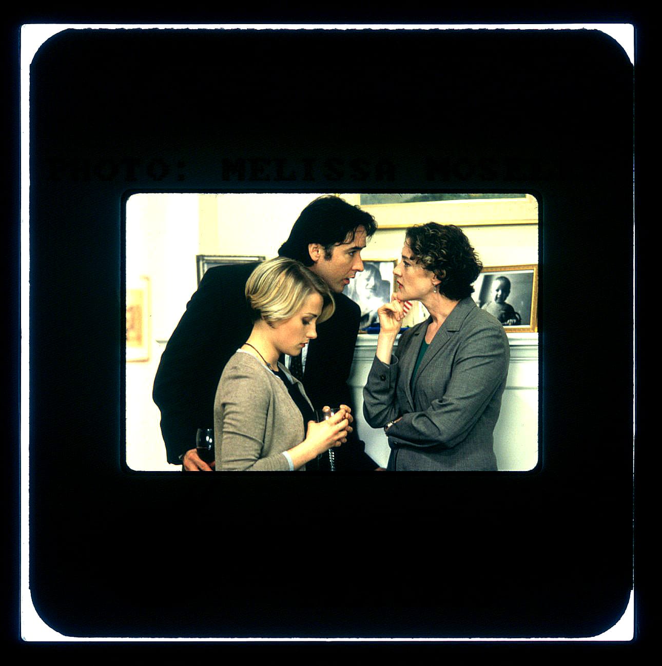 Color Publicity Slide of three people. A man in black on left leans in close towards the face of a woman in suit coat with chin resting on right hand. Another woman is in front of the man, head down so his head is fully visible above.