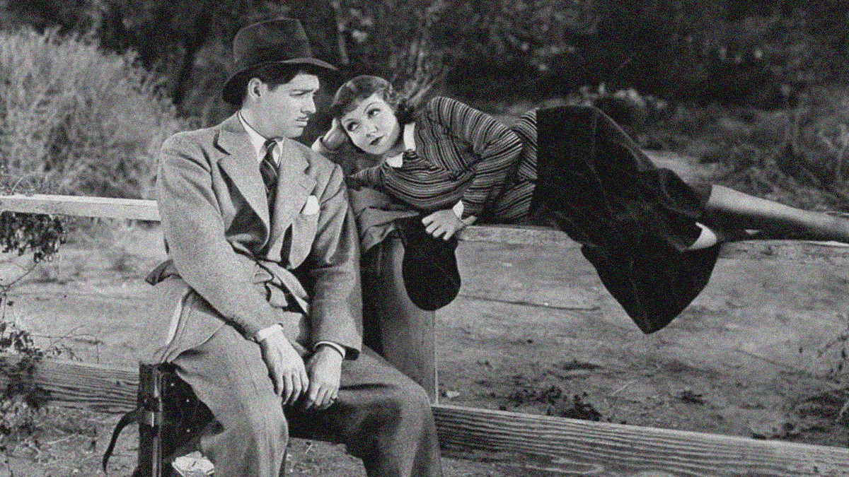 A man in suit and hat sits dejectedly on his suitcase while a woman lying on a wooden fence slat smiles at him.