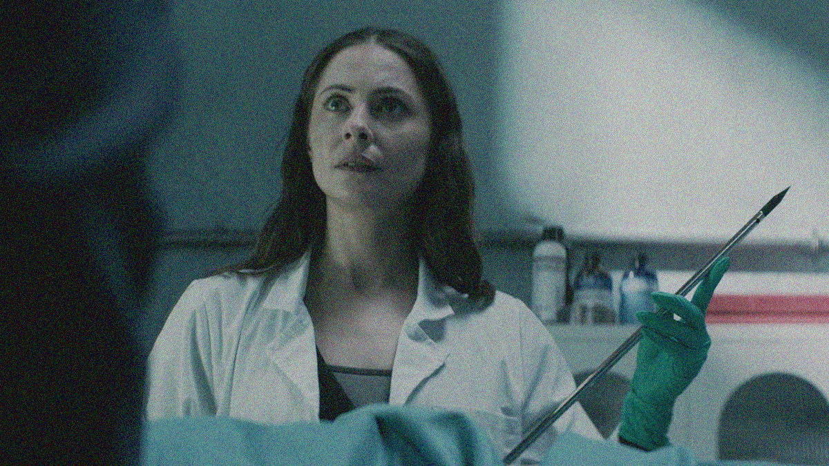 A woman in lab coat is seen from chest up looking off-screen upwards left. She holds an embalming needle injector.