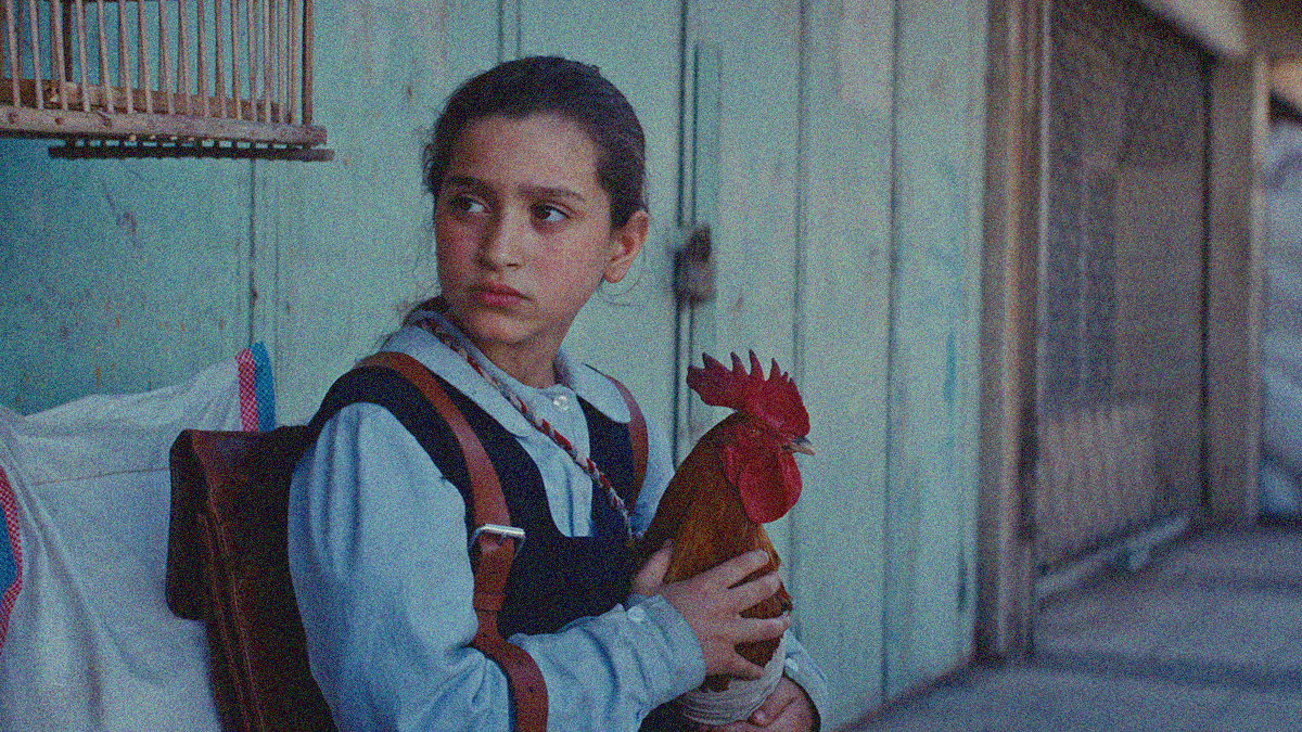 A young girl in school uniform sits alone outside a sparse building with a rooster in her hands.